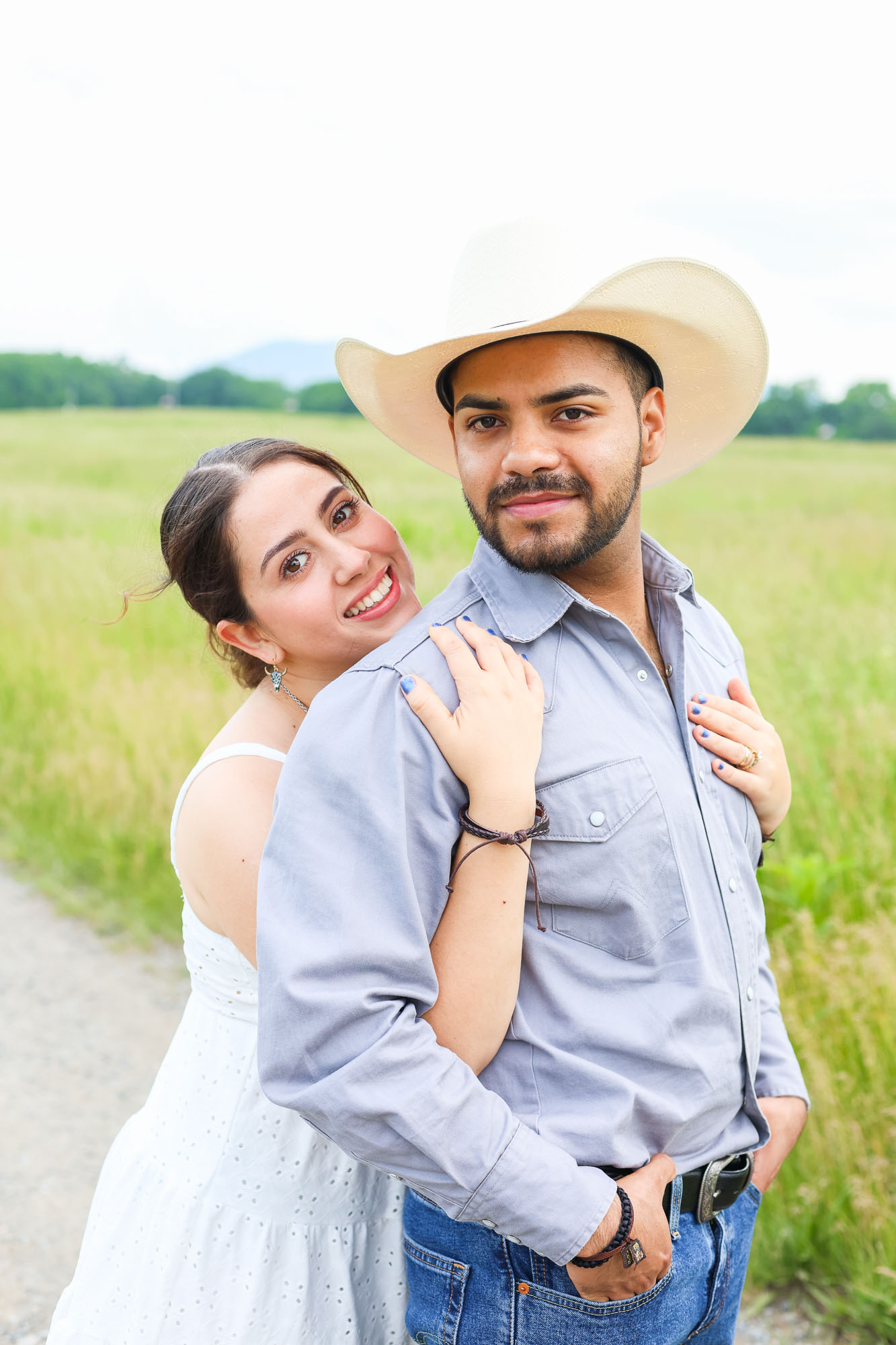 A couple stands together in a grassy field, with the woman playfully leaning on the man from behind. The woman wears a white dress, while the man has on a gray shirt and a wide-brimmed white cowboy hat.