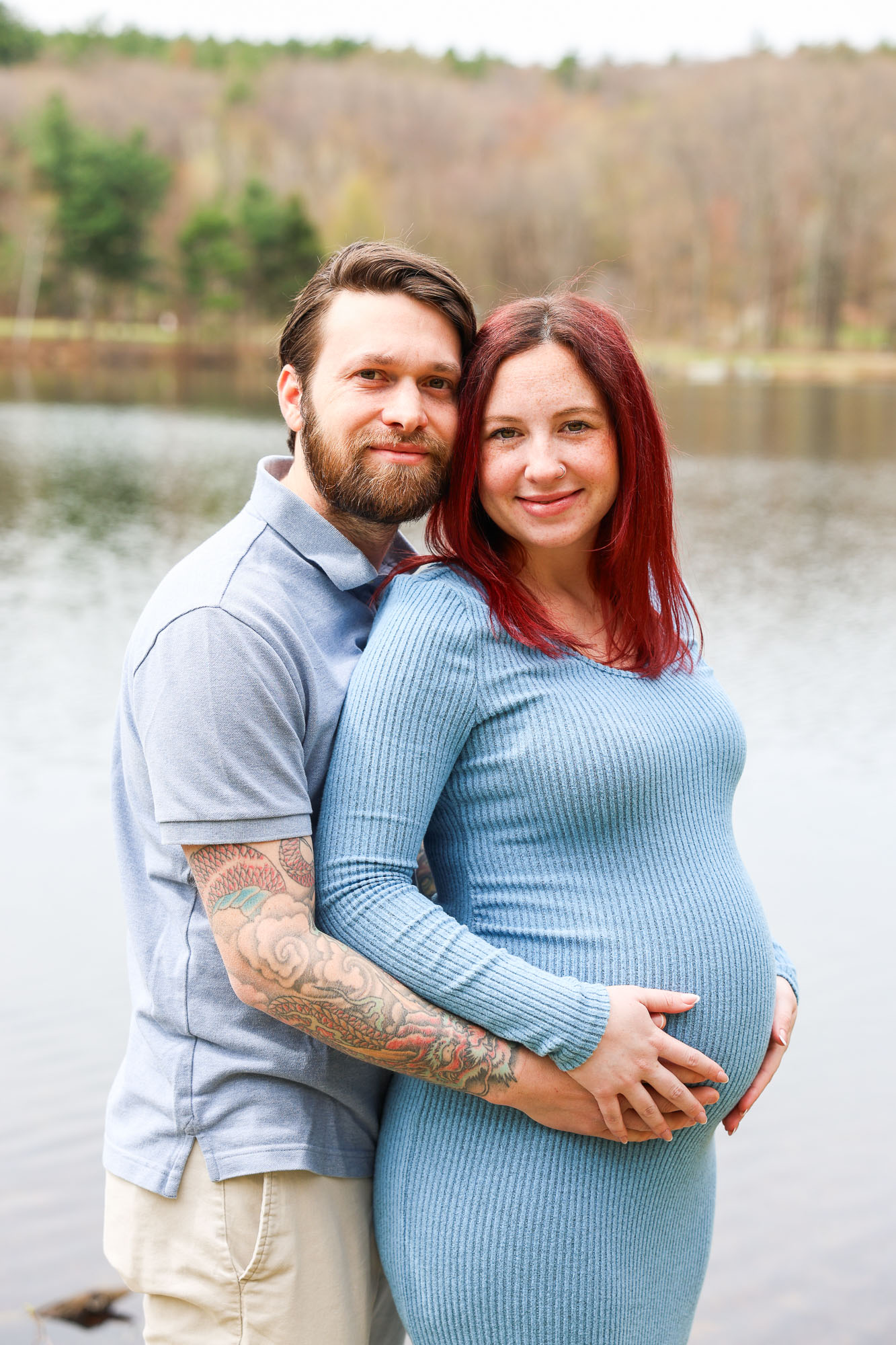 A couple embracing by a serene lake, with the woman showing her baby bump. The man has a beard and tattoos, and the woman has long red hair. They are both smiling.