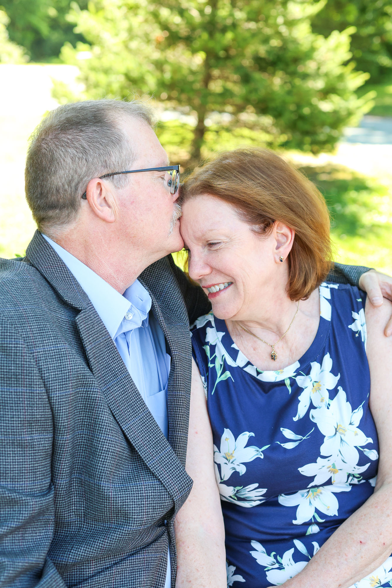 A couple smiling affectionately, with the man kissing the woman's forehead; they are seated outdoors surrounded by greenery.