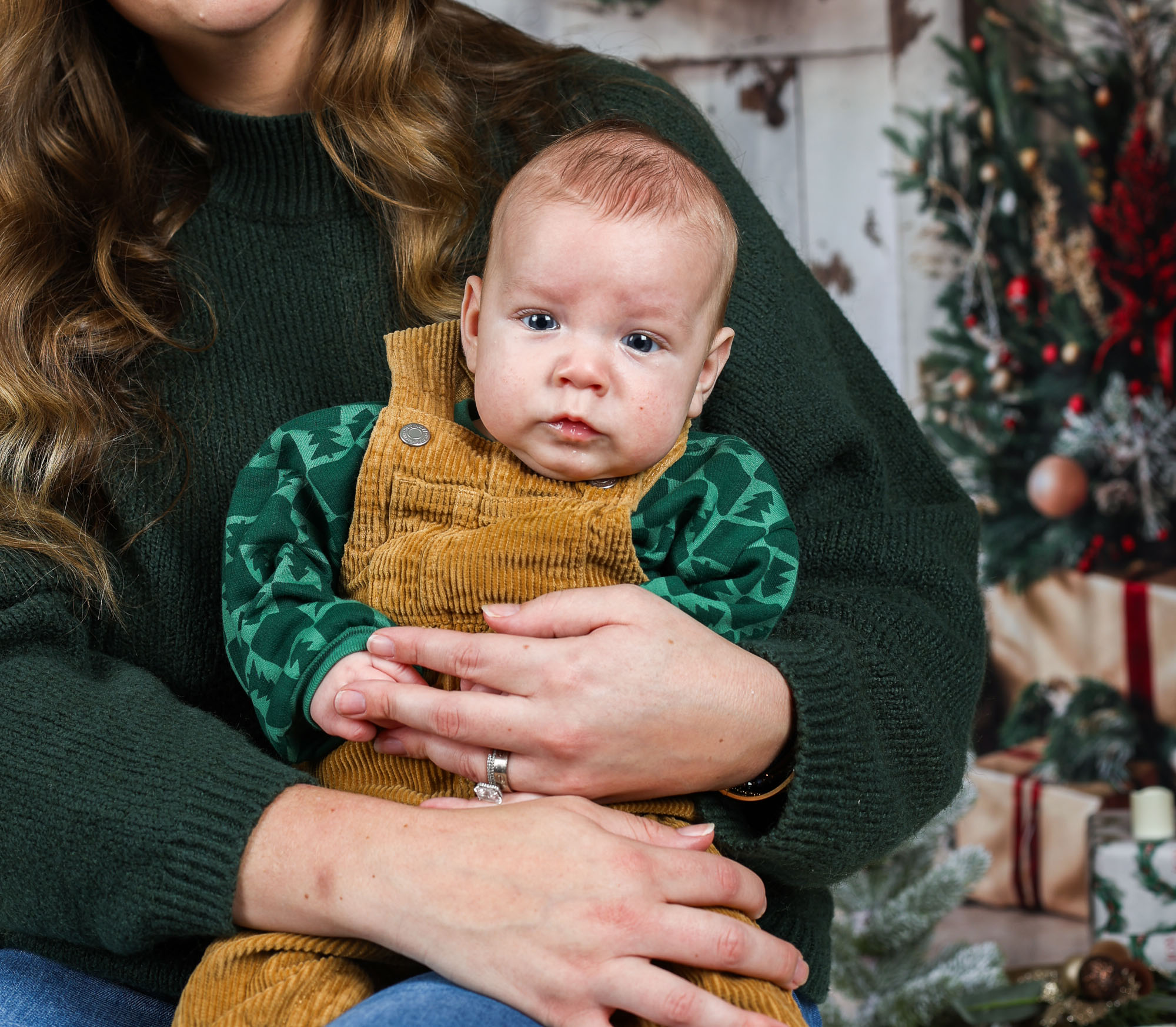 A close-up of a baby in a brown corduroy overall, sitting on a woman's lap, with a green patterned long-sleeve shirt. The background features a festive Christmas tree and wrapped presents.