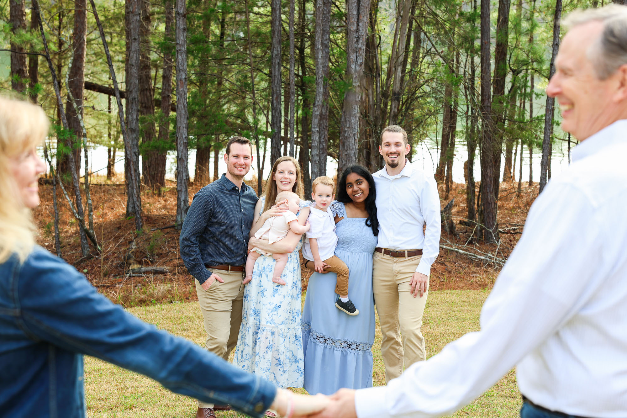 A group of six people standing in a forest clearing, smiling and posing for the camera. One woman is holding a baby, while a young child stands next to her. The group includes a mix of adults in casual attire. Tall trees and a calm lake are visible in the background.