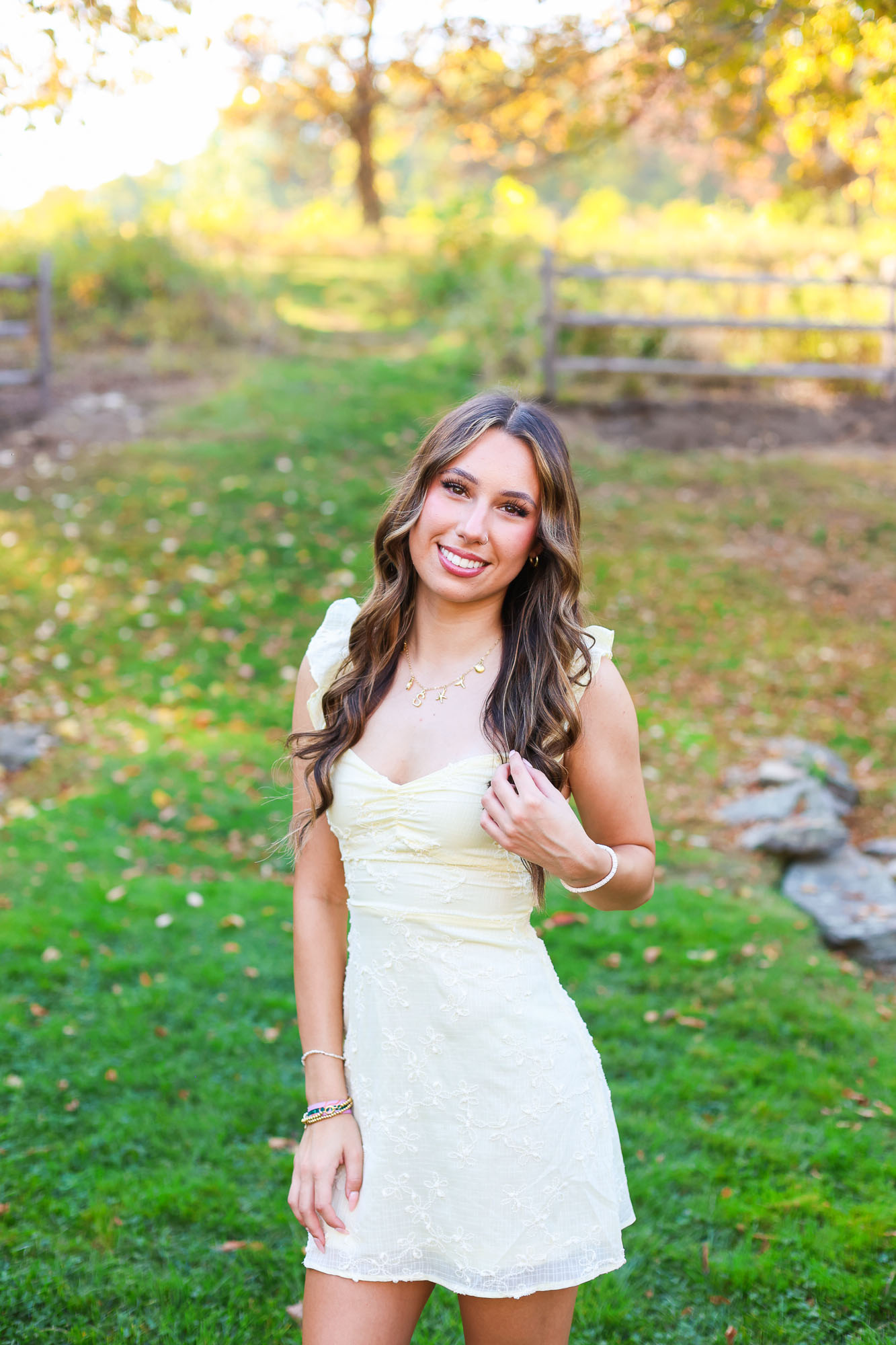A young woman smiling while standing on green grass, wearing a light yellow dress with floral detailing and accessories, set against a background of trees and a wooden fence.