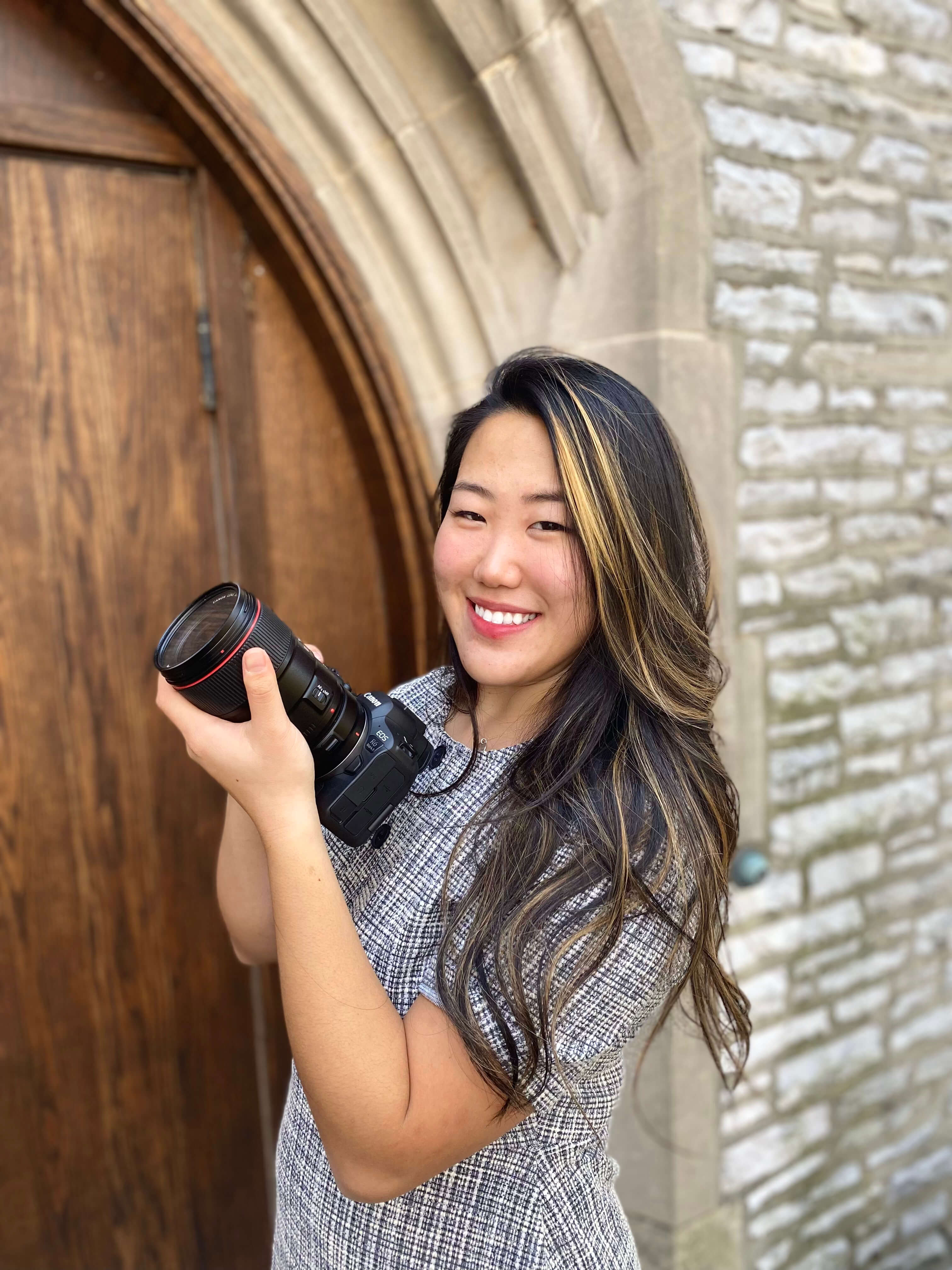 A smiling young woman holding a camera, standing in front of a wooden door with stone walls in the background.