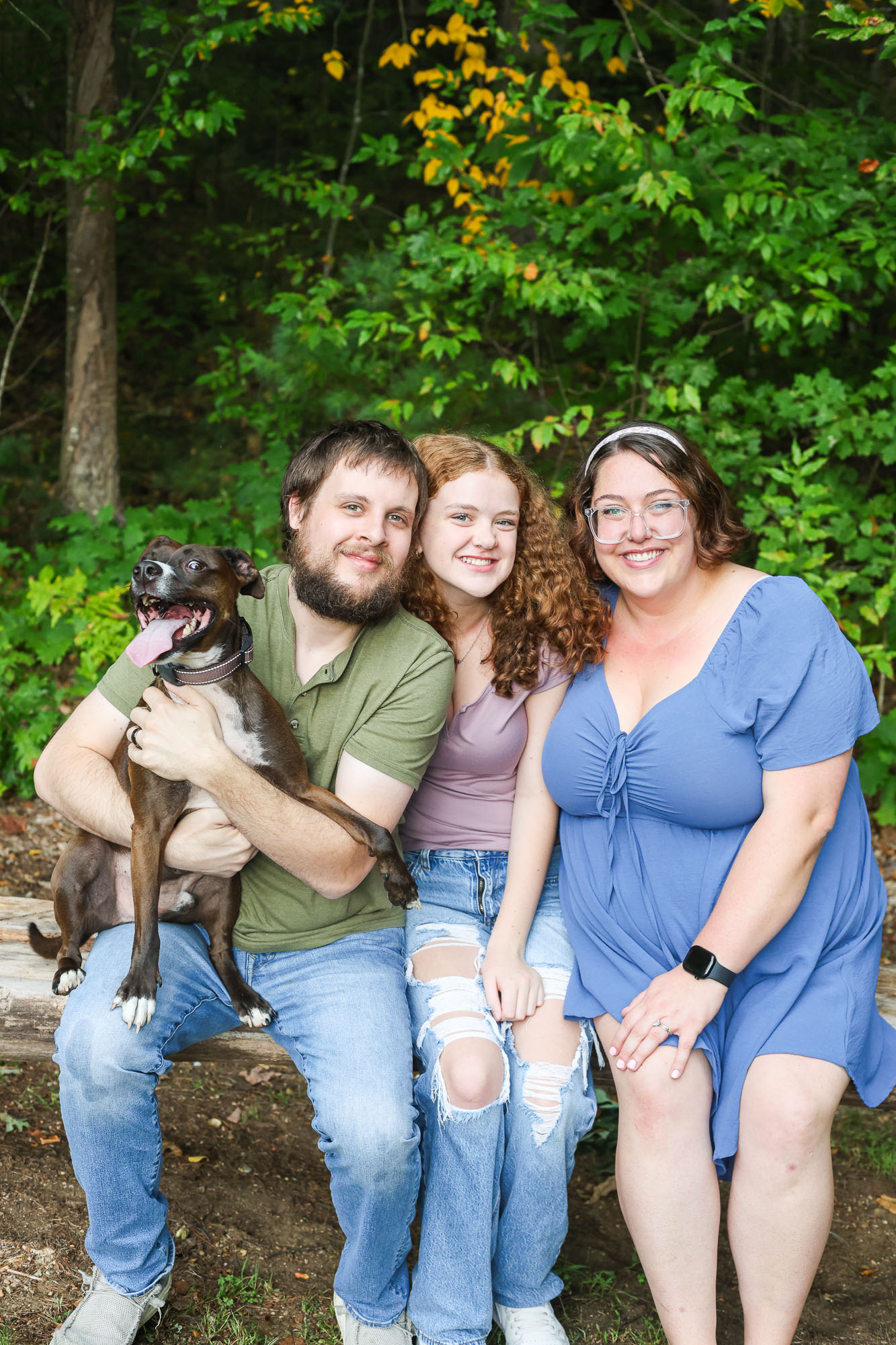 A family portrait featuring three individuals sitting on a log in a natural setting with green foliage. A man is holding a small dog in his arms, while a teenage girl and a woman sit next to him, all smiling at the camera.